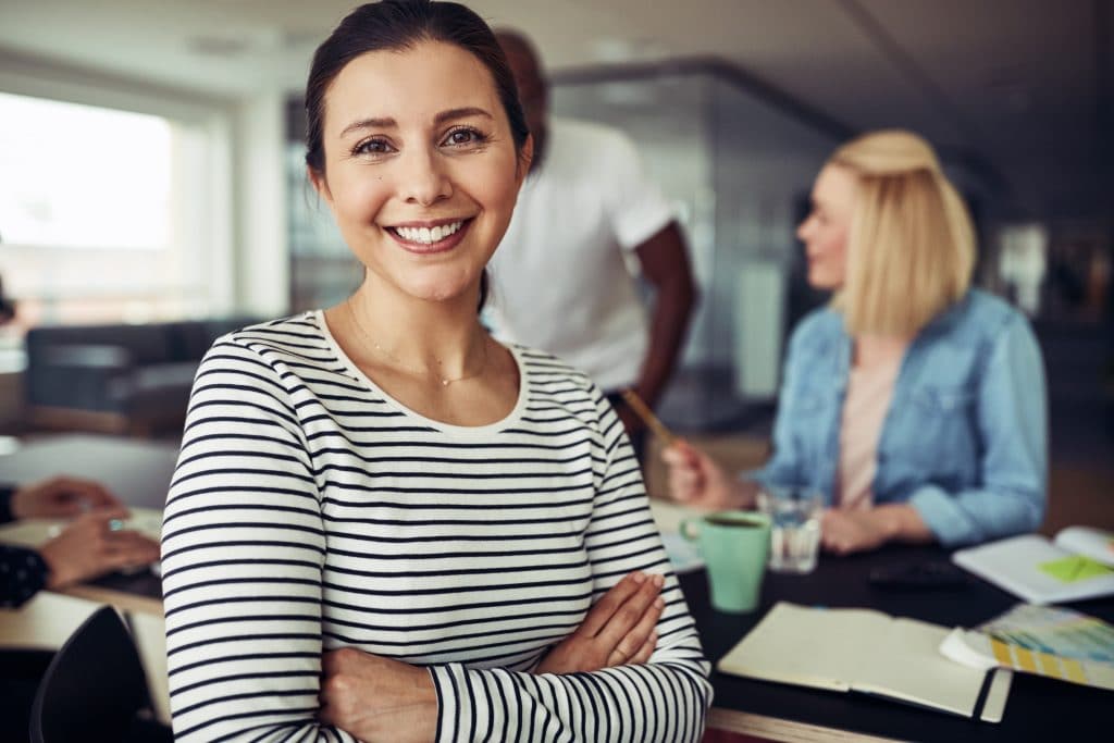 Jeune femme d'affaires souriante et assise les bras croisés à une table de bureau avec des collègues parlant ensemble en arrière-plan